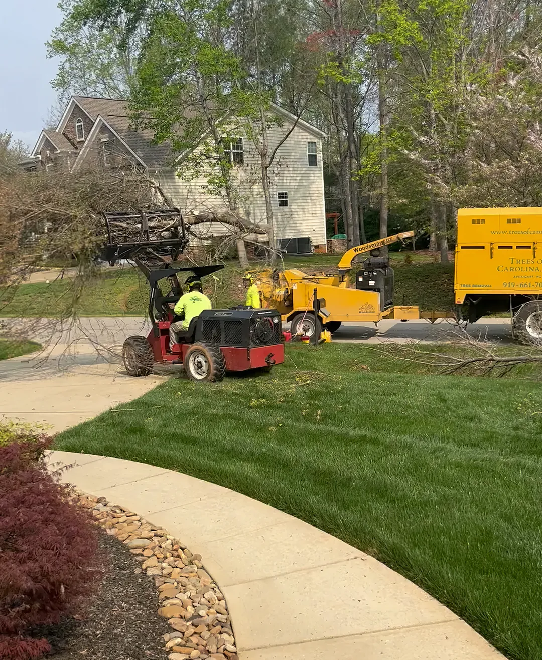 Red grapple loader carrying brush toward a chipper and yellow chip truck beside a curving front walk